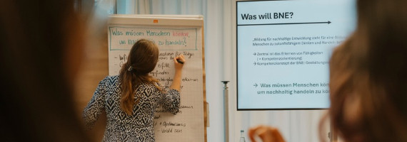 Eine Person mit langen, braunen Haaren beschriftet eine Flipchart vor ihr. Rechts daneben ist ein Bildschirm aufgebaut. Text: Was ist BNE?