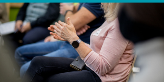 Klatschende Hände einer sitzenden Person, im Hintergrund mehr Sitzende.